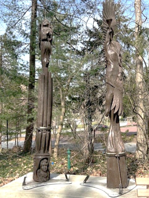 Close view of both sculptures on their new concrete pads at the Chautauqua grounds, forest green trees behind them